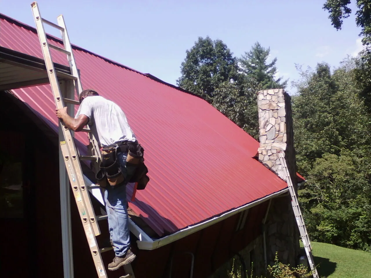 Roofer on a ladder beside a red metal roof and stone chimney — Soffit & Fascia Repair by Roofing Brothers in Ahoskie